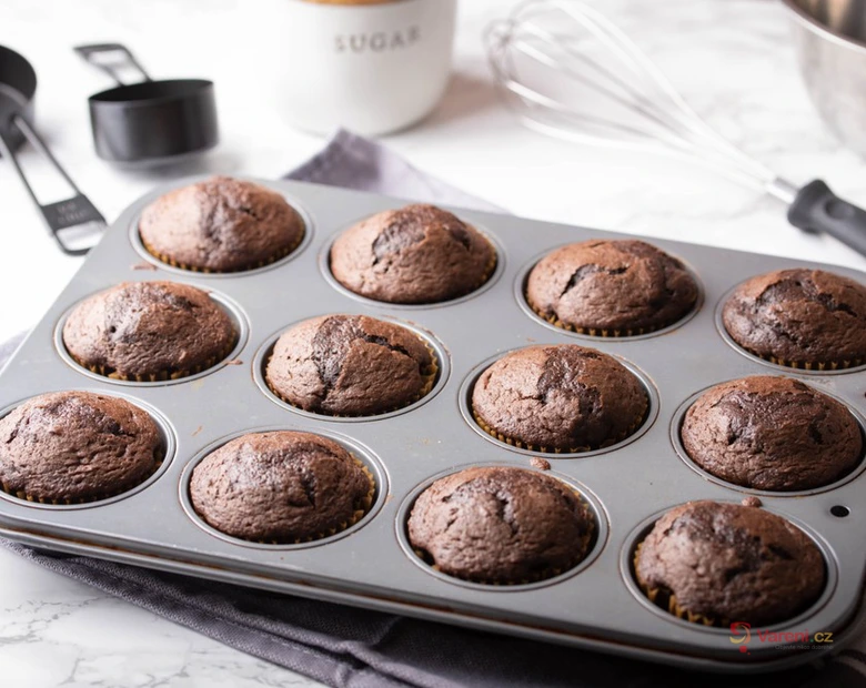 Čerstvé čokoládové muffiny v pekáči s kuchyňskými nástroji.