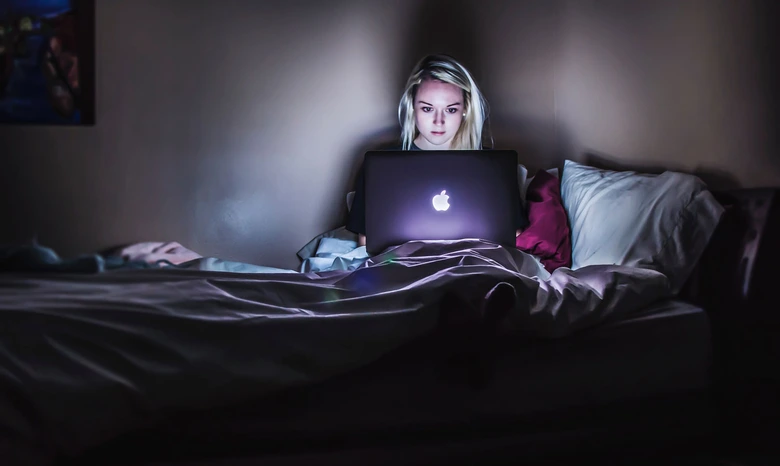Žena sedí v posteli s otevřeným notebookem ve tmě.