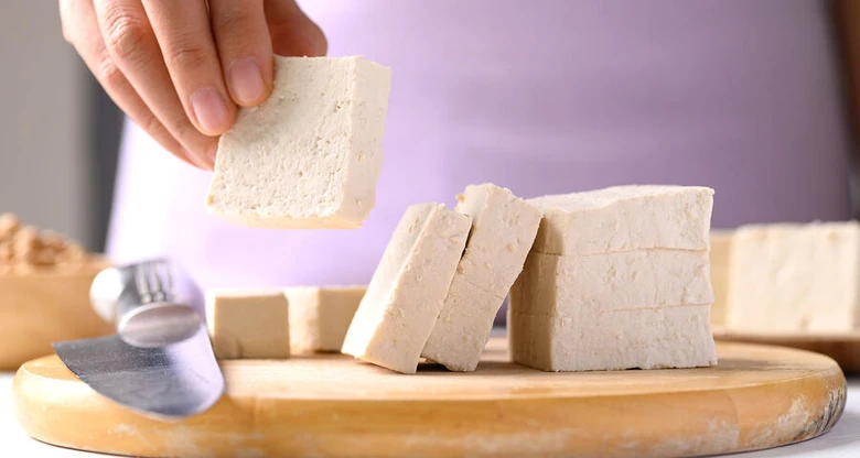Ruce drží kus čerstvého tofu nad dřevěnou prkénkem.