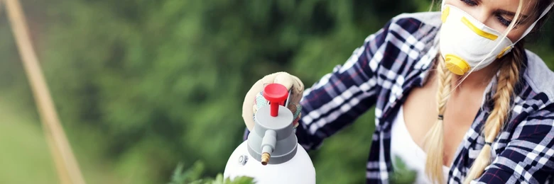Žena v ochranné masce postříká rostliny pesticidem ručním rozprašovačem.