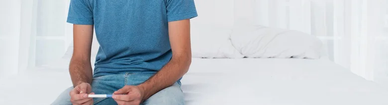 Muž sedící na posteli drží v ruce diagnostickou tyčinku.