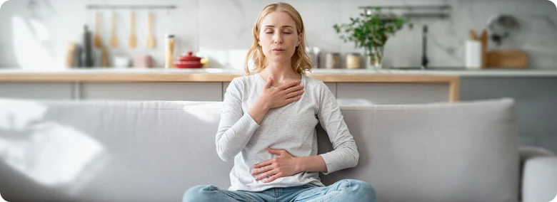 Žena sedí na gauči v kuchyni s dlaněmi na hrudi při meditaci pro úzkost.