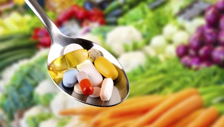 Lžíce s vitamíny a doplňky stravy před pozadím zeleniny.