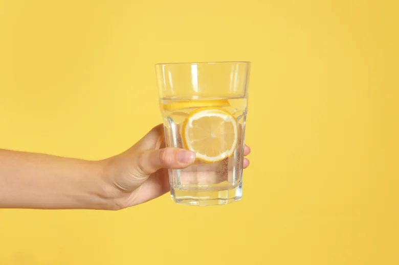 Ruka drží sklenici vody s citronem na žlutém pozadí.
