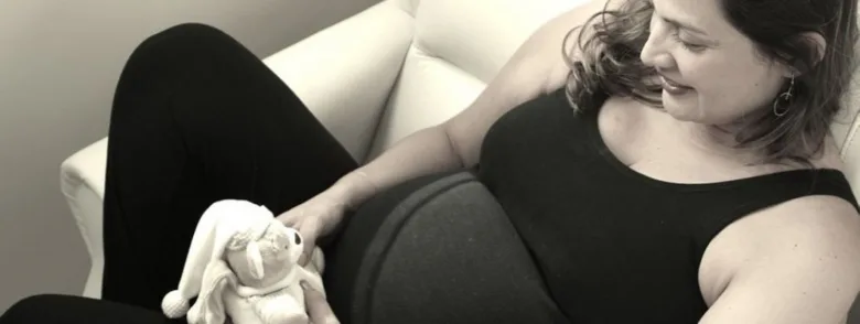 Těhotná žena sedí na gauči s hračkou u břicha.