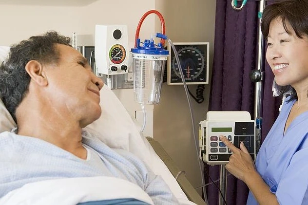Muž v nemocničním lůžku s lékařkou upravující infuzní čerpadlo.