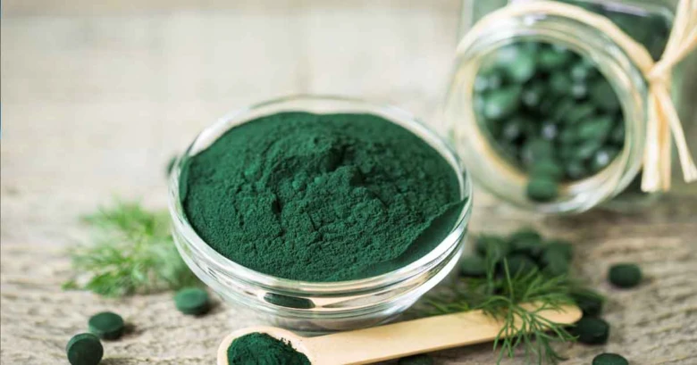 Skleněná miska s práškem z řasy Spirulina vedle skla tablet.