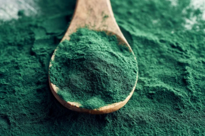 Dřevěná lžička s jasně zelenou práškem spiruliny na pozadí.