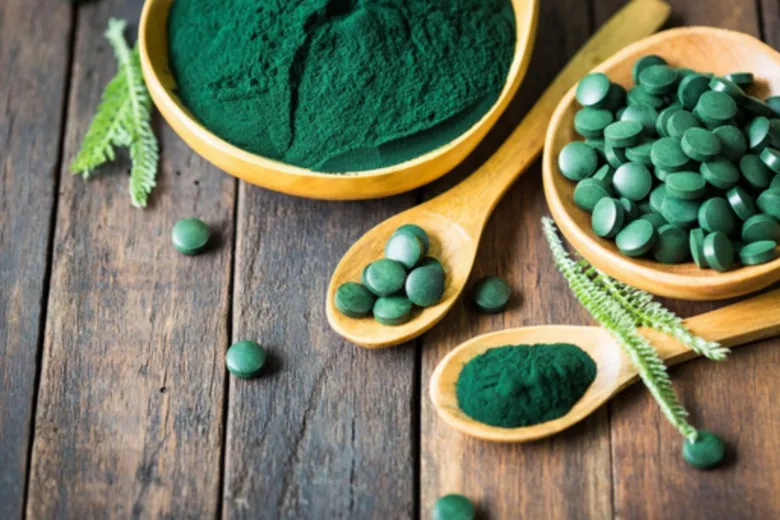 Dřevěná deska s miskou prášku z řasy Spirulina a lžičkami tablet.