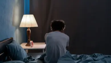 Osoba sedí na posteli v tmavé místnosti s rozsvícenou lampou.