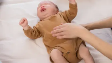 Pláčající miminko v hnědém bodýtku na posteli s rukou dospělého.