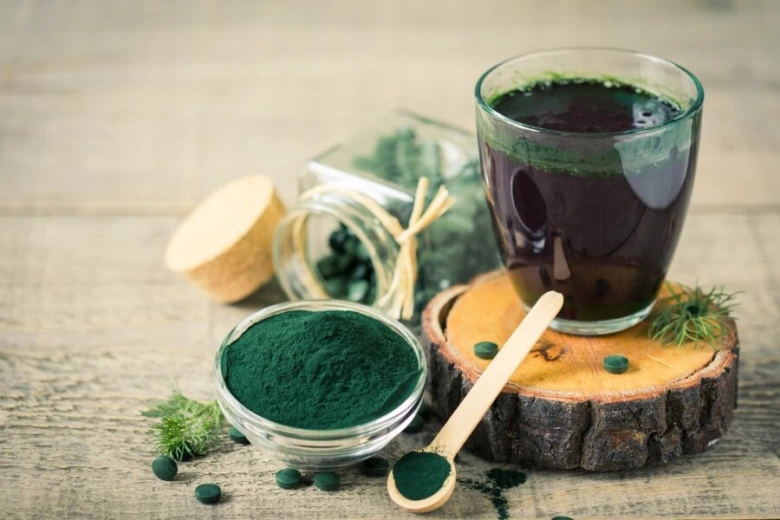 Sklenice s tekutou spirulinou vedle prášku a kapslí na dřevěném stole.