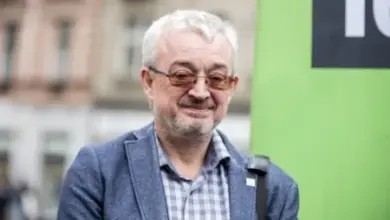 Muž s brýlemi před zelenou tabulí na ulici.