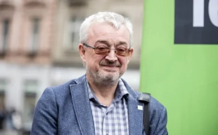 Muž s brýlemi před zelenou tabulí na ulici.