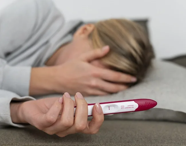 Žena drží pozitivní těhotenský test, zatímco odpočívá na polštář.