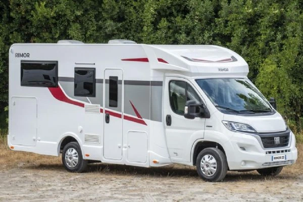 Bílý obytný vůz typu karavan zaparkovaný před zelenými stromy.