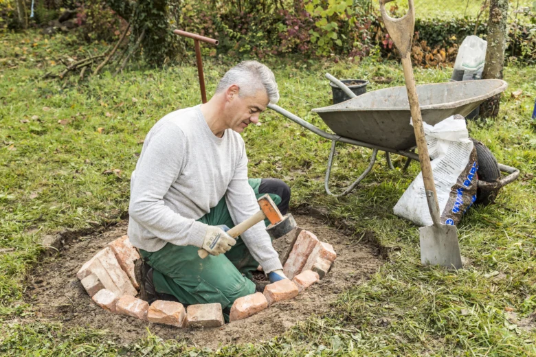 Muž staví cihlové ohniště v zahradě klečící na zemi.