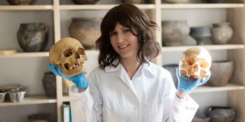 Žena v laboratorním plášti drží dva lidské lebky.