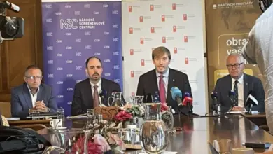 Tři muži sedí za stolem na tiskové konferenci s mikrofony a kamerami.