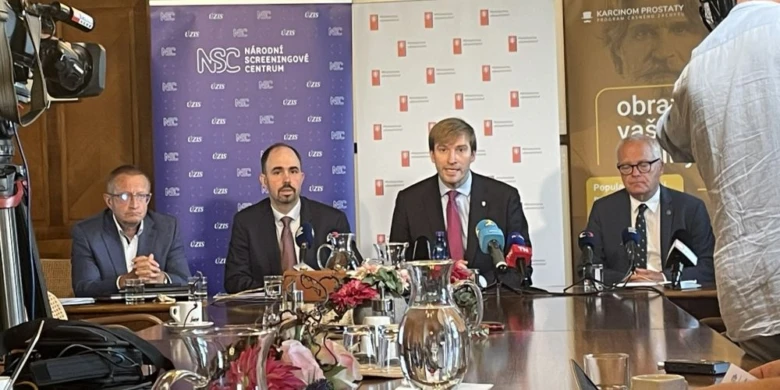 Tři muži sedí za stolem na tiskové konferenci s mikrofony a kamerami.