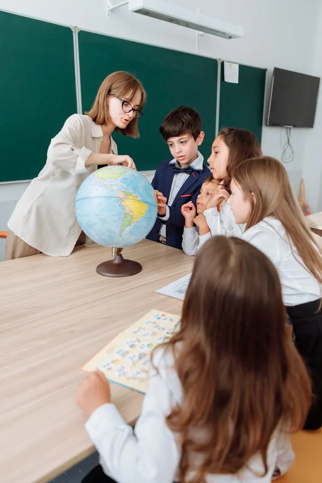 Učitelka ukazuje na zeměpisnou kouli před skupinou dětí v učebně.