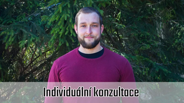Muž v červeném tričku stojí venku před jehličnatými stromy.