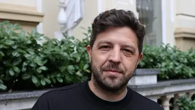 Muž s vousy v černém tričku před zelenými keři.